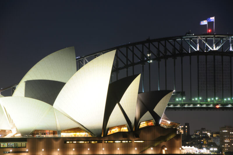 Illuminated Sydney Opera House shell roofs before Sydney Harbour Bridge steel arch at night