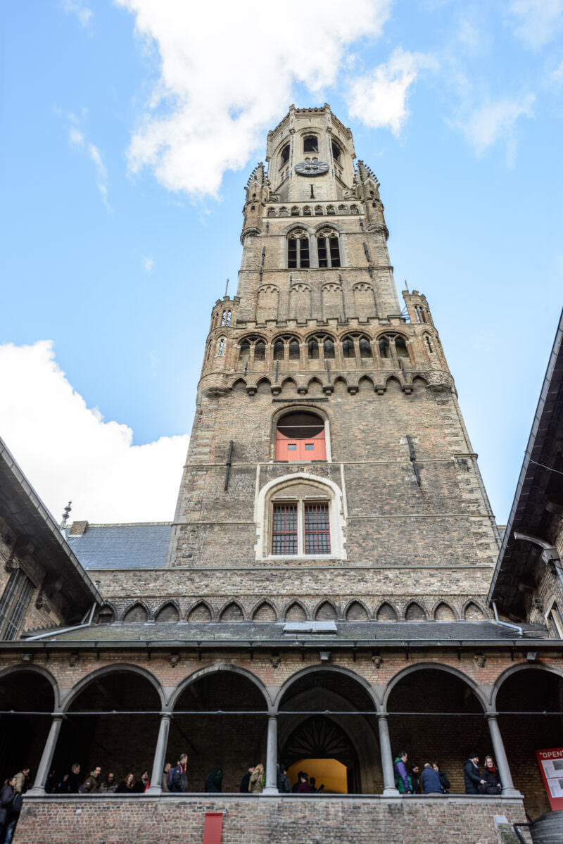 Belfry of Bruges rear facade rising from an internal courtyard, showing brick lower stages and sandstone octagonal lantern against cloudy sky