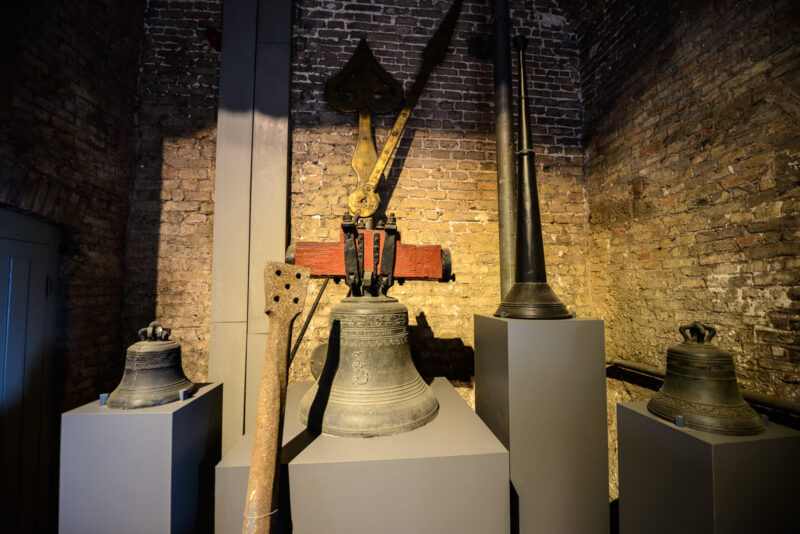 Display of historic carillon bells from the Belfry of Bruges, including large bronze bells with wooden yokes and clamps against illuminated brick wall