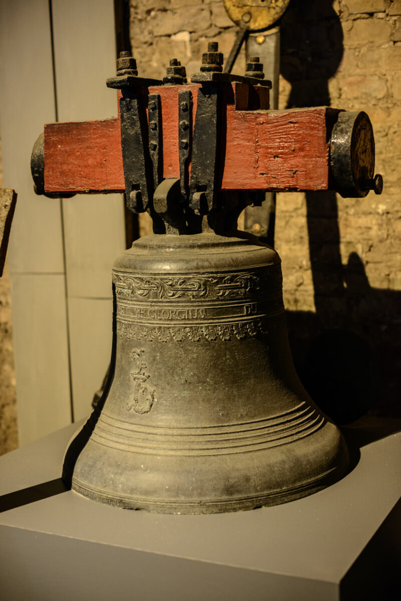 Historic bronze bell with decorative bands and inscription, mounted on a wooden yoke with iron clamps against aged brick wall