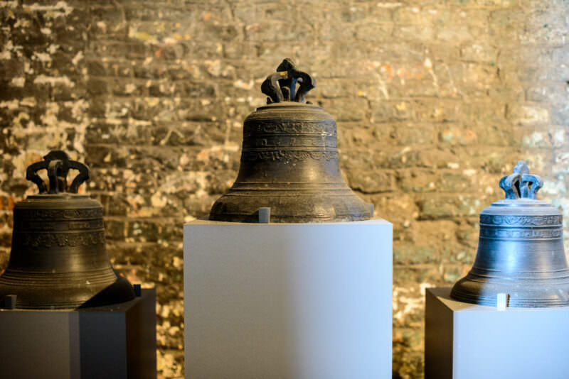 Three bronze bells with decorative crowns displayed on white pedestals against illuminated golden brick wall in the Belfry exhibition