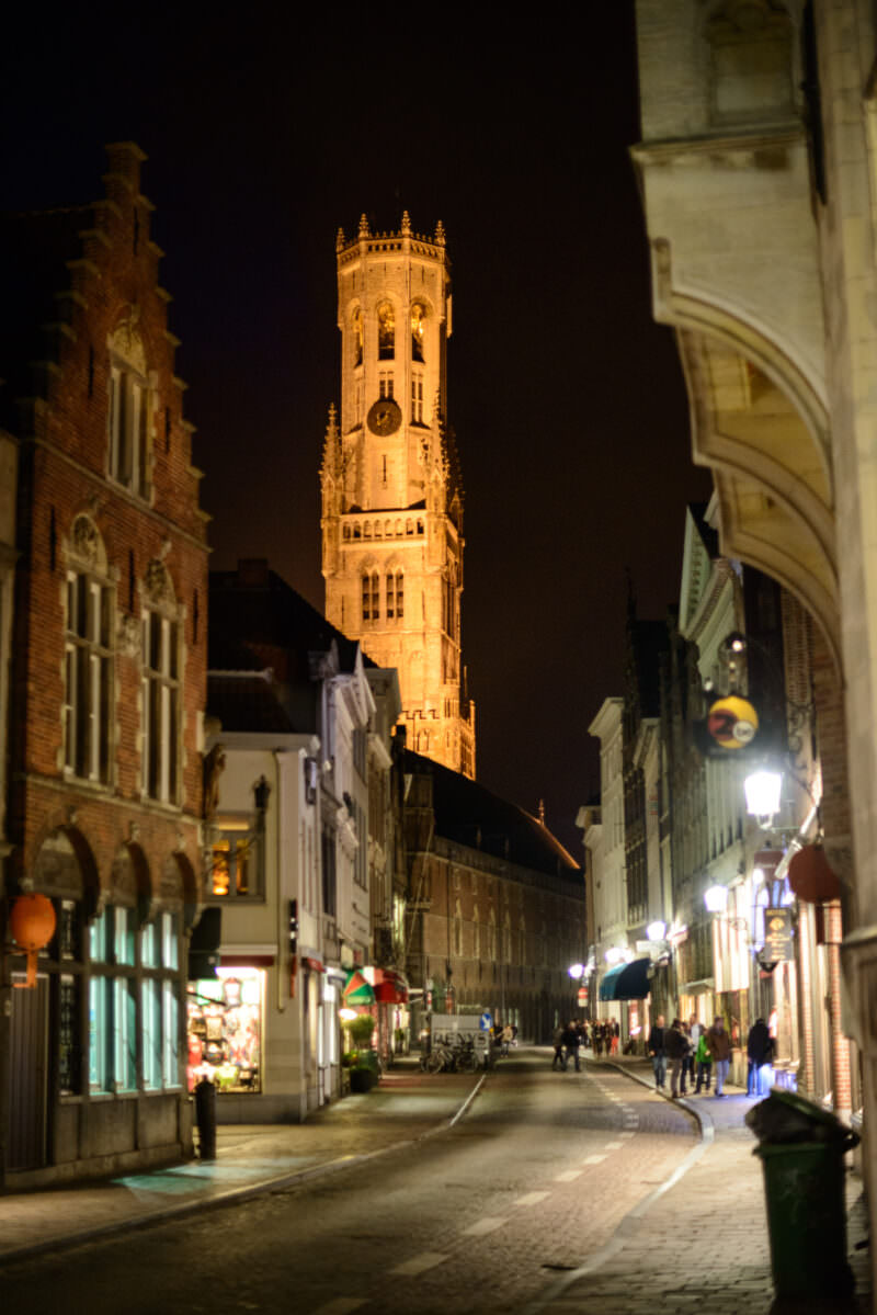 Belfry of Bruges illuminated at night, its octagonal lantern glowing at the end of a narrow medieval street framed by historic buildings