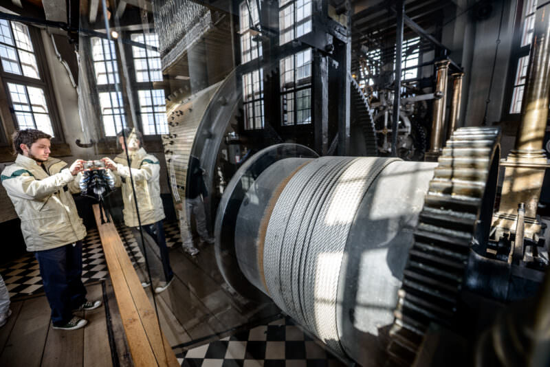 Large steel-wrapped drum and pendulum mechanism of the Belfry carillon, with a visitor photographing the machinery in the checkered-floor chamber