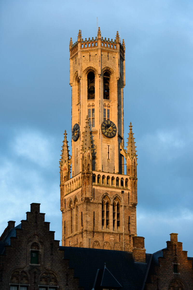 Belfry of Bruges in morning sunlight, its octagonal crown and clock faces illuminated above the Markt, framed by historic gabled buildings