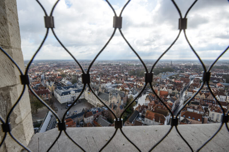 Bruges medieval cityscape viewed through decorative iron grillwork from atop the Belfry, showing red-tiled roofs and the Markt below
