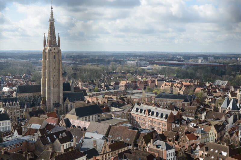 Church of Our Lady spire and Bruges rooftops spreading to the horizon, viewed from the Belfry observation level under cloudy sky