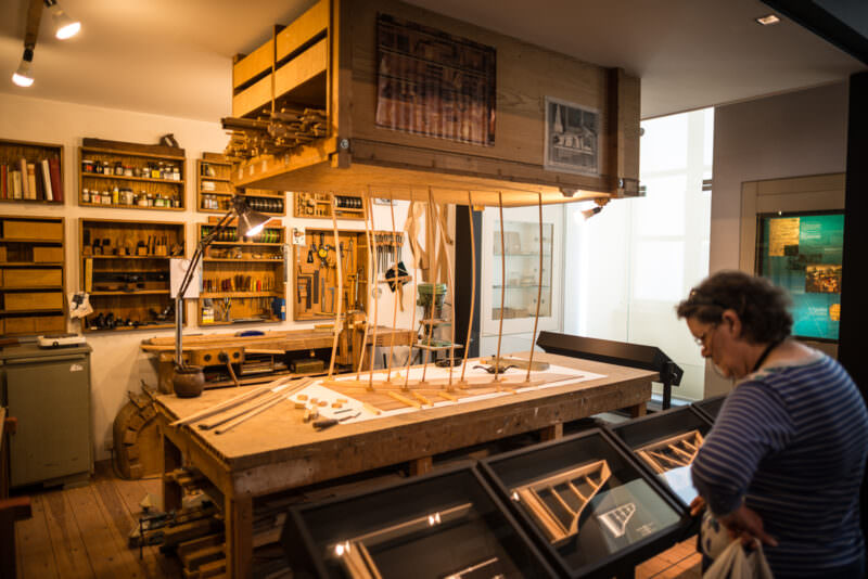 Reconstructed piano workshop with wooden go-bars clamped vertically above workbench, tools lining walls behind glass