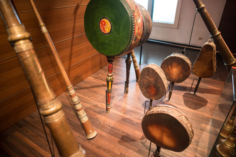 Brass telescoping horns and painted ceremonial drums with green leather heads mounted on wooden display stands