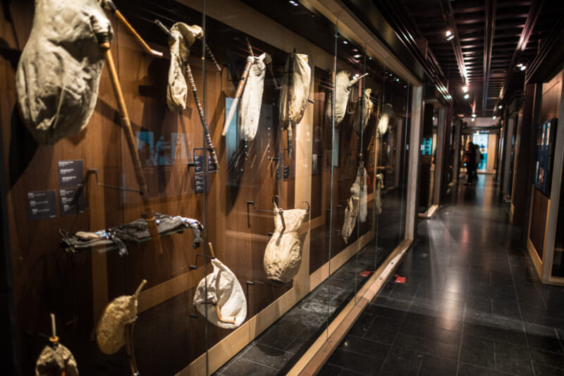 Dozens of bagpipes with animal-hide reservoirs and wooden drones hanging along glass-walled museum corridor