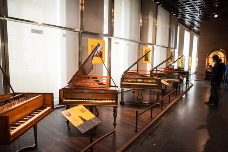 Gallery of early pianos and fortepianos in wooden frames, floor-to-ceiling windows casting natural light across polished floor