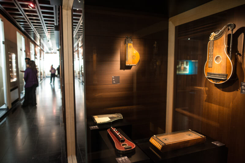 Chord zithers with button mechanisms and fretted autoharp bodies displayed along glass corridor under ceiling lights