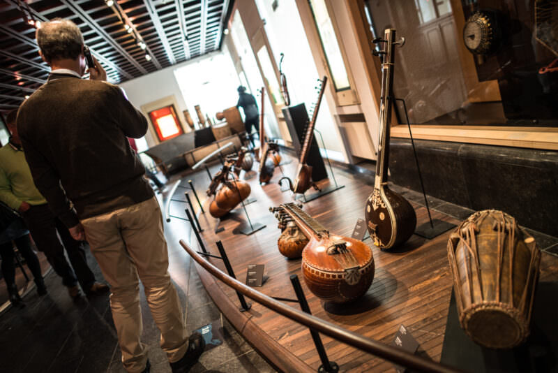 Tanpura and mridangam drum on tiered display platform, visitor with headphones approaching in modern gallery space