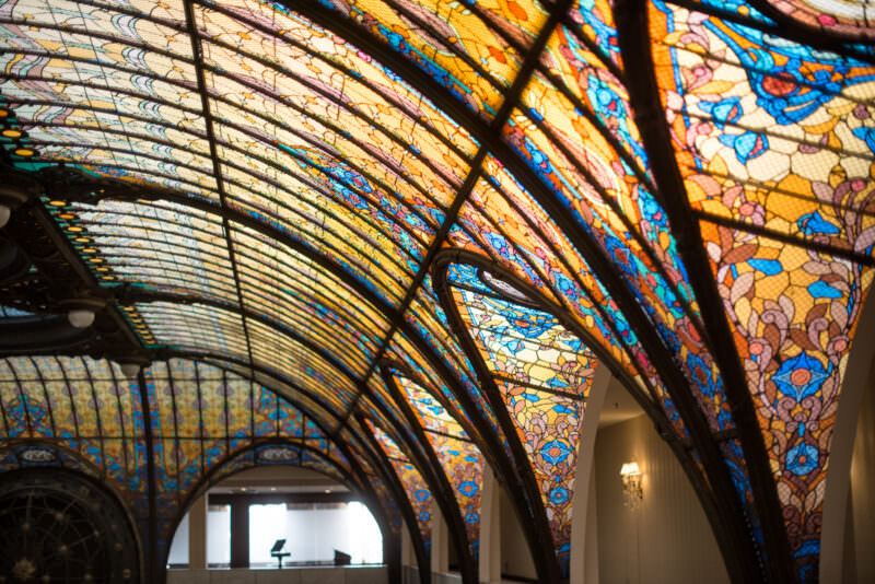 Arched stained glass section of Gran Hotel Ciudad de Mexico ceiling showing curving ironwork and floral motifs in amber and blue