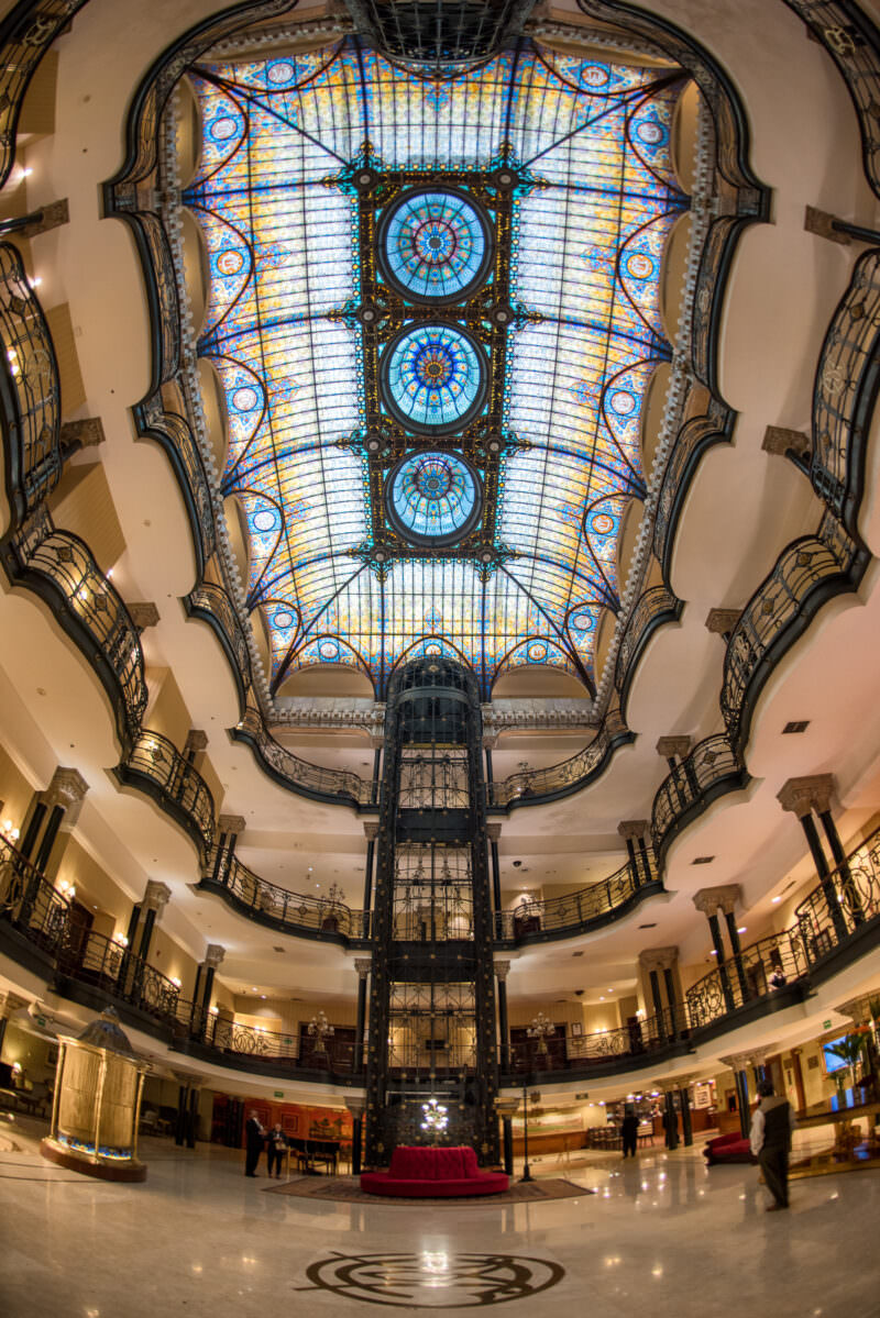 Upward view of Gran Hotel Ciudad de Mexico atrium showing three medallions centered in the stained glass vault with ornate ironwork