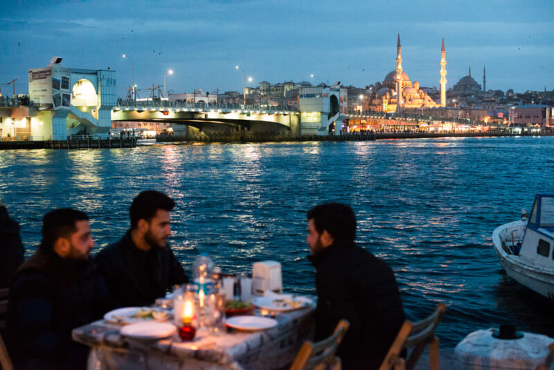 Waterside diners at Karaköy face the illuminated New Mosque and Galata Bridge across the Golden Horn at dusk