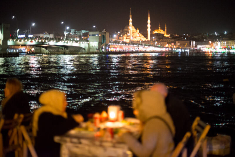 Karaköy waterfront diners with the New Mosque, Galata Bridge, and Süleymaniye Mosque skyline across the Golden Horn
