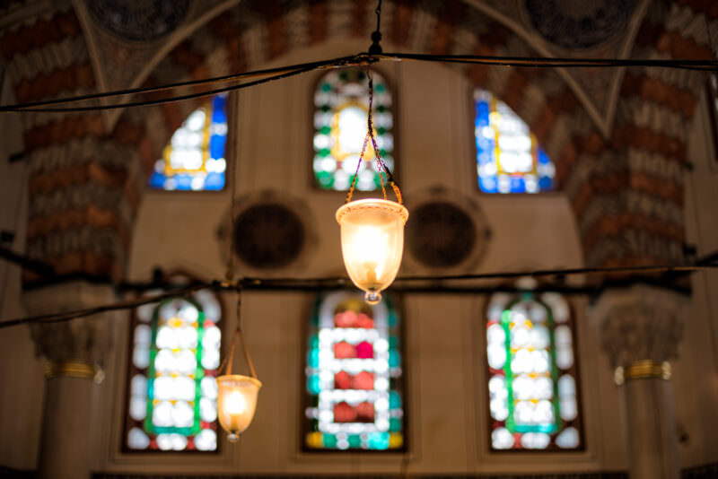 Colorful stained-glass windows glowing above hanging oil lamps inside the Tomb of Sultan Murad III
