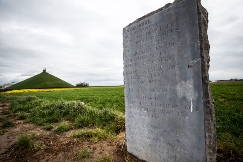 Stone memorial slab with French inscription in foreground, Lion's Mound and yellow rapeseed field behind