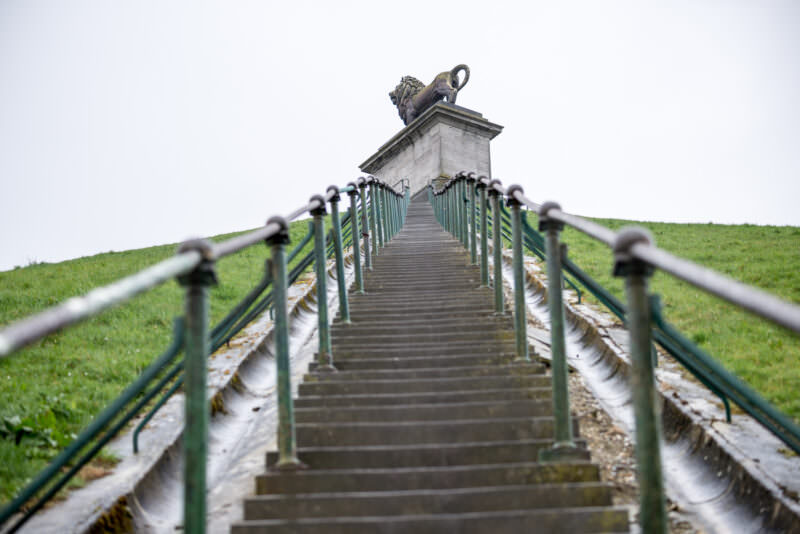 Steep staircase ascending the Lion's Mound toward pedestal and lion statue, chain-link railing on both sides
