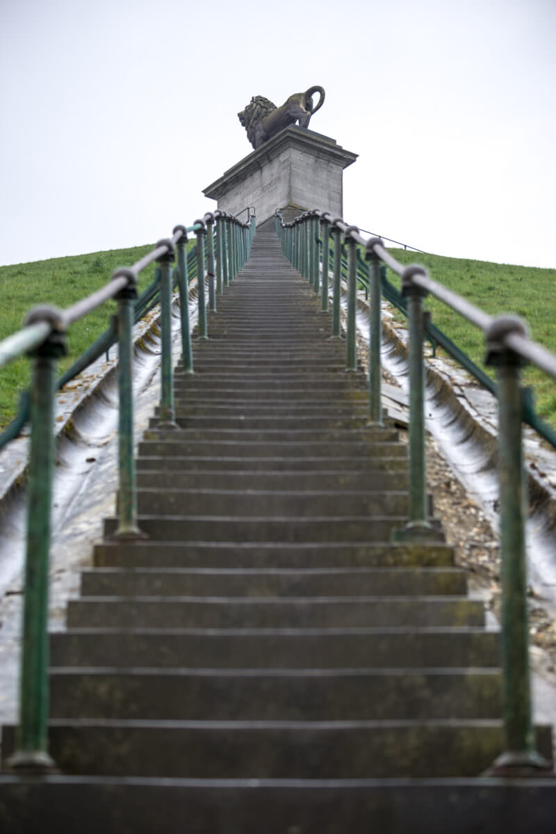 Lion statue atop pedestal with Roman numeral inscription, viewed from staircase with chain-link railing in foreground