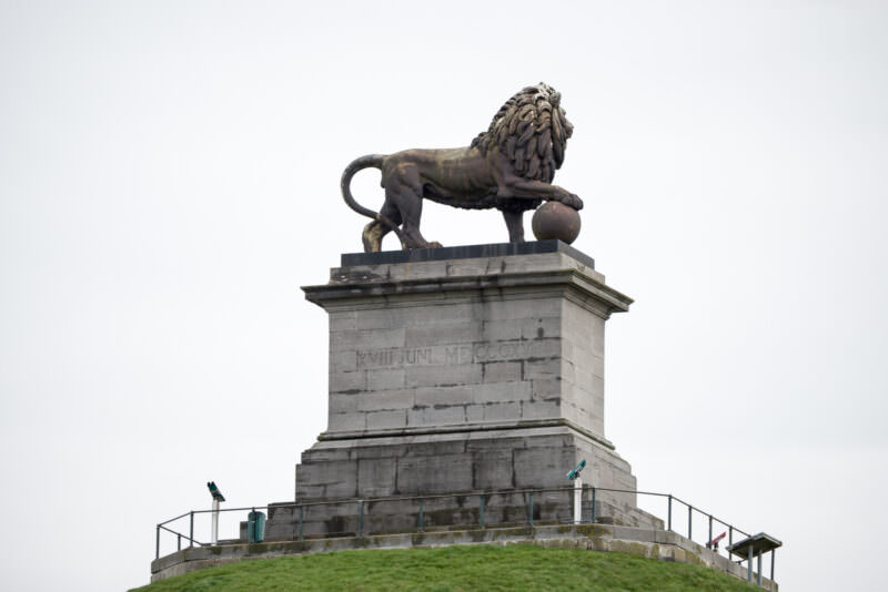 Lion statue with paw on globe atop weathered stone pedestal with Roman numeral inscription, railing visible below