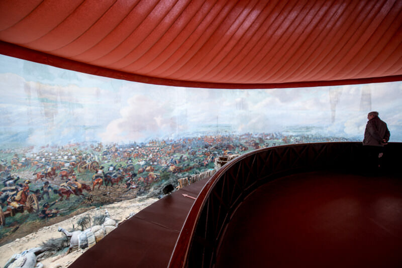 Interior of the Panorama rotunda showing painted cavalry charge under fabric canopy, with a single visitor silhouetted at railing