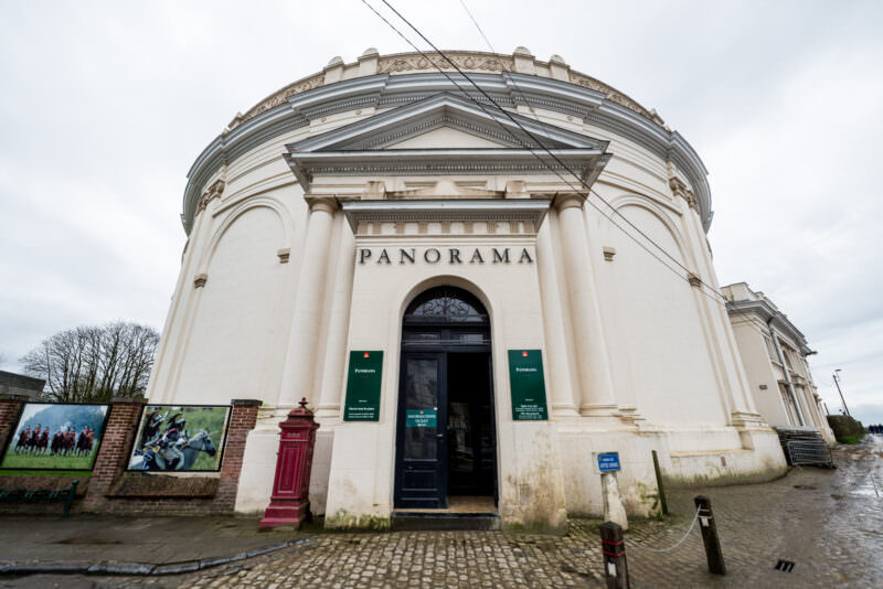White neoclassical Panorama rotunda with pedimented entrance spelling PANORAMA, green banners flanking dark doorway