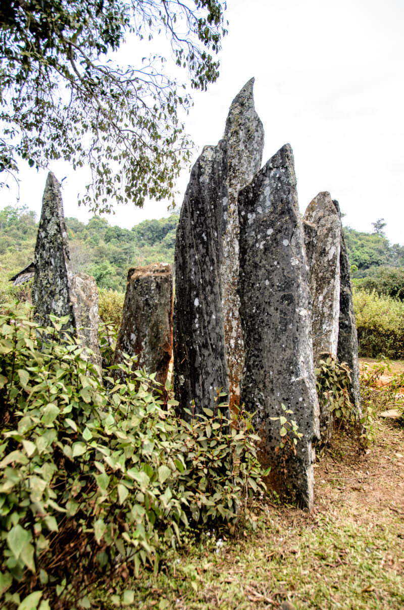 Cluster of tall Hintang menhirs covered in lichen standing against an overcast sky, surrounded by low vegetation and forested hills