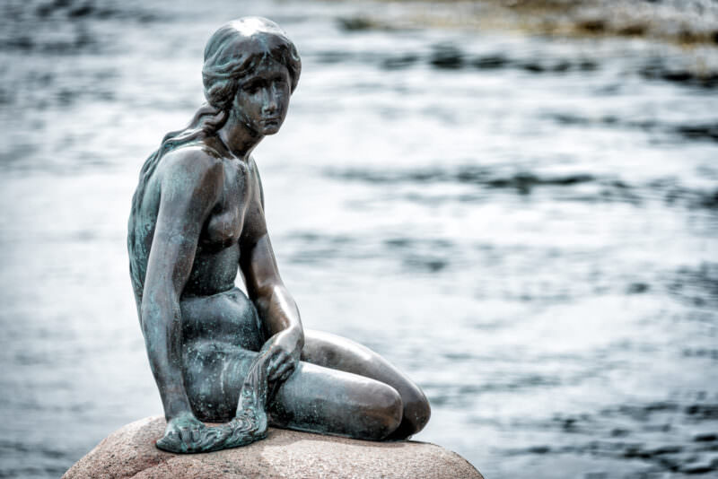 The Little Mermaid statue in profile, showing her braided hair and weathered patina on bronze surface against harbour backdrop