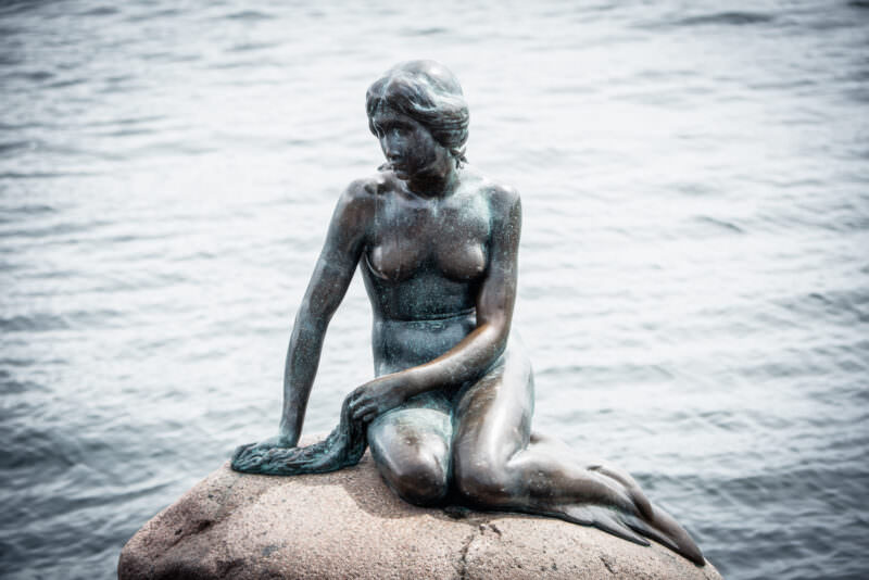 The Little Mermaid statue in Copenhagen, weathered bronze figure with fish tail seated on granite boulder against grey harbour water