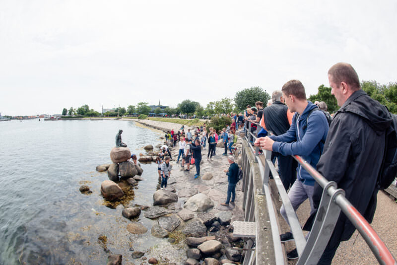 The Little Mermaid statue on its boulder surrounded by visitors on rocky shoreline with crowds gathering along the promenade railing