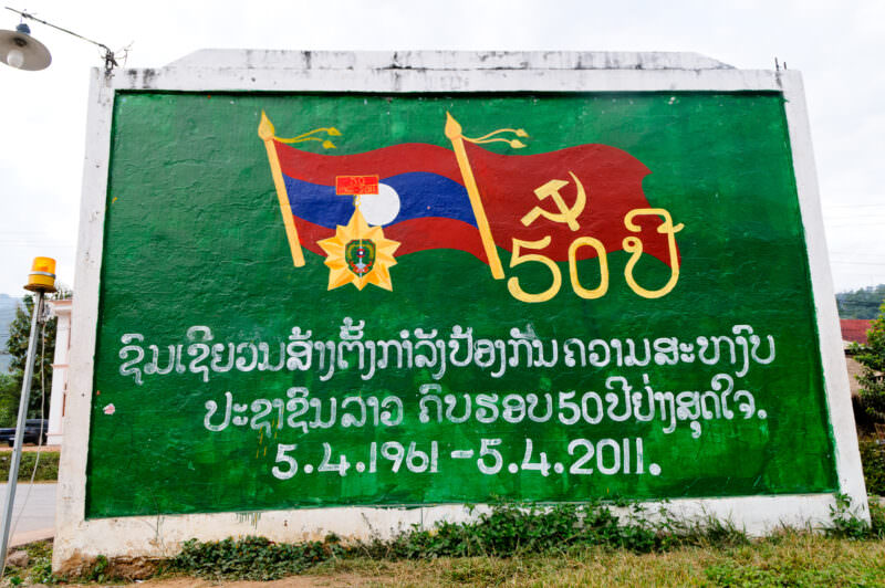 Green painted billboard showing the national flag and hammer-and-sickle emblem with Lao script in white lettering