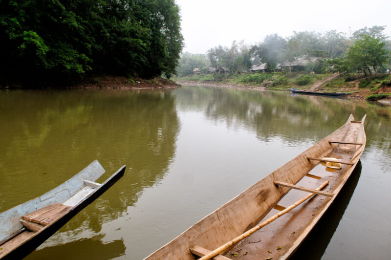 Two wooden longboats on the muddy banks of the Nam Tha River, mist obscuring far shore and dwellings