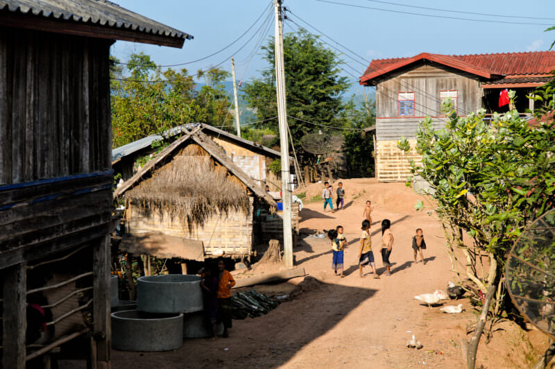 Children gathered on a dirt road among wooden and bamboo stilt houses with metal and thatch roofs, mountains behind