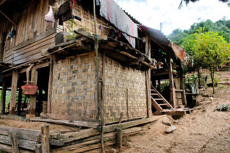 Chickens foraging beneath a stilted house with woven bamboo walls and wooden ladder, hillside and vegetation behind