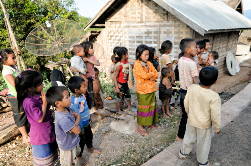 Children standing in a dirt clearing beside woven bamboo homes and a large mesh satellite dish, mountains behind