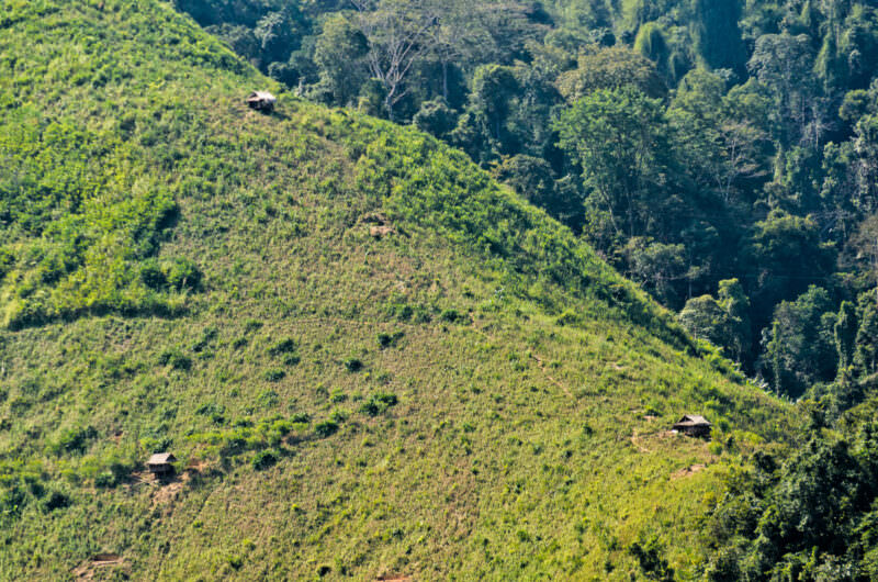 Small bamboo huts on a steep cleared mountainside, surrounded by green vegetation and forested peaks