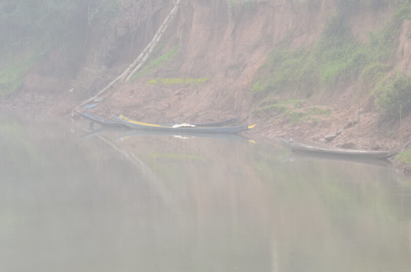 Two wooden longboats partially obscured by morning mist on the Nam Tha River, steep earthen banks visible