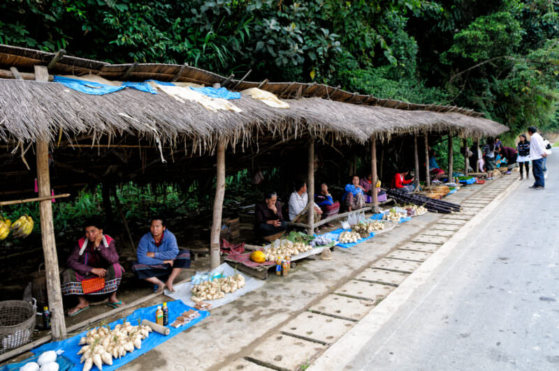 Vendors under a long thatched shelter displaying produce on tarps along a paved roadside, forested hills behind