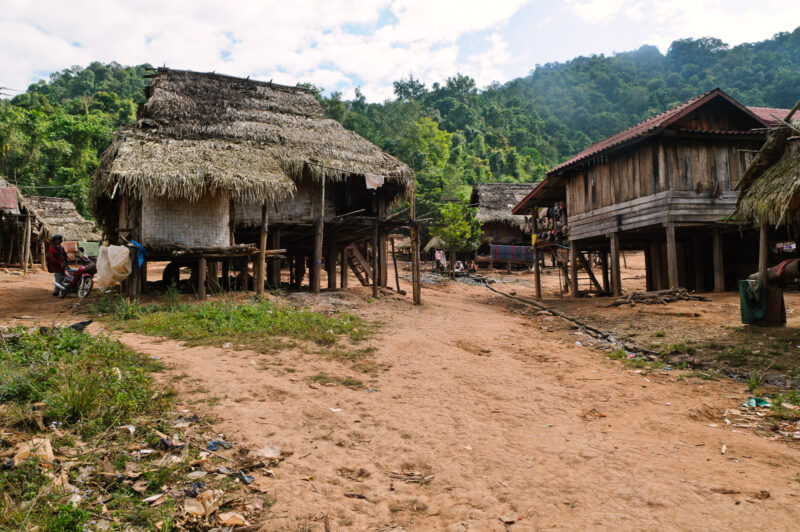 Wooden and bamboo stilt houses in Lakkhamma Village with thatched roofs, foreground dirt road and forested hills behind