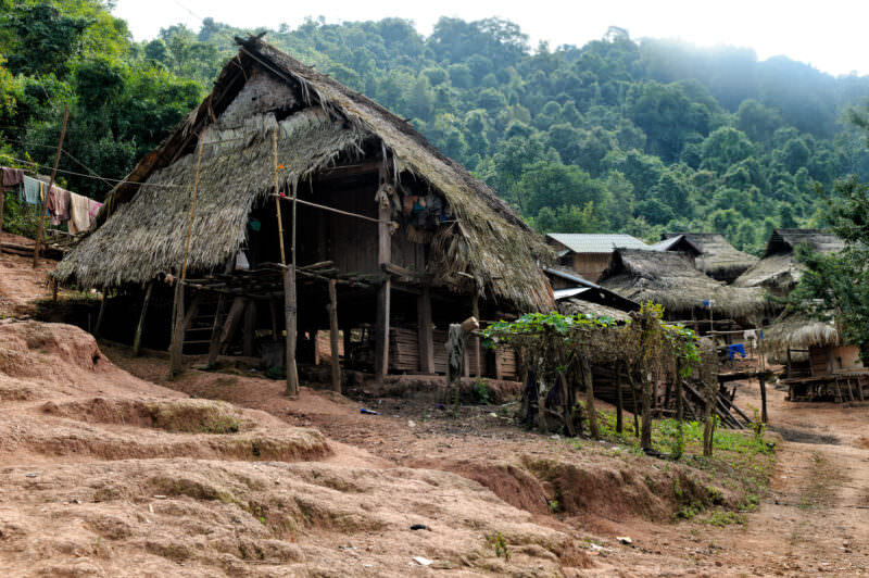 Thatched-roof stilt houses lining a dirt hillside in Lakkhamma Village, misty forested hills in background