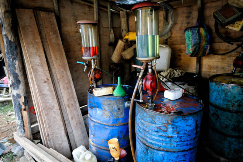 Hand-cranked fuel pumps with glass cylinders atop blue 55-gallon drums inside a wooden shack with plank walls