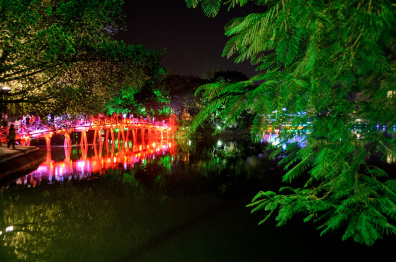 Pedestrians on the glowing red Huc Bridge framed by a green fern branch over Hoan Kiem Lake