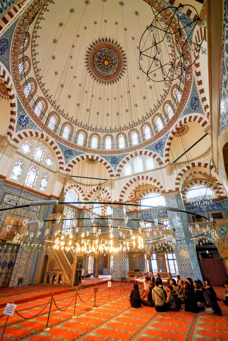 Rustem Pasha Mosque's prayer hall with tiled octagonal walls, a domed ceiling, and visitors seated on the red carpet