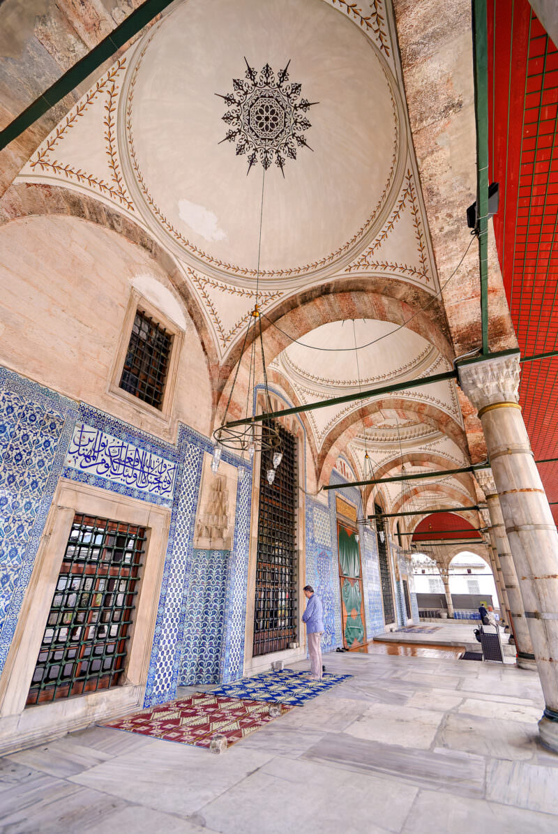 Man standing in prayer beside Iznik-tiled walls along Rustem Pasha Mosque's entrance cloister