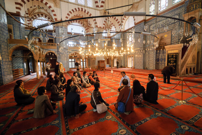Tour group seated on the carpet inside Rustem Pasha Mosque's tiled prayer hall beneath a ring of hanging lamps