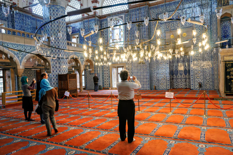 Visitors photographing Rustem Pasha Mosque's floor-to-ceiling Iznik-tiled prayer hall beneath a chandelier