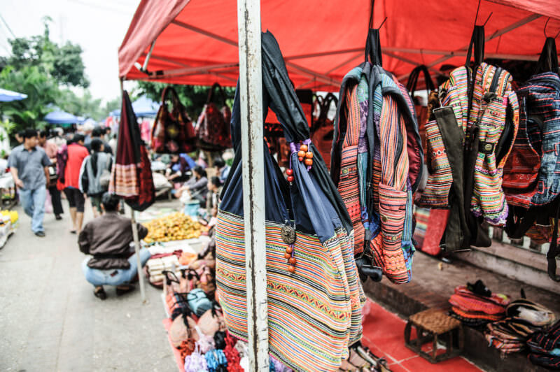 Handwoven striped bags in red, blue, and cream hanging from a market stall under a red canopy