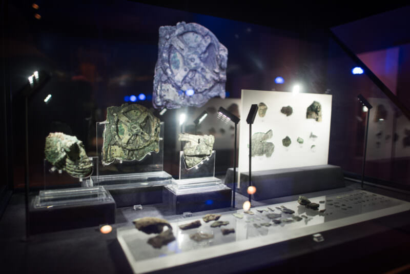 Antikythera Mechanism fragments in transparent display stands, corroded bronze pieces with visible gearing in a museum case