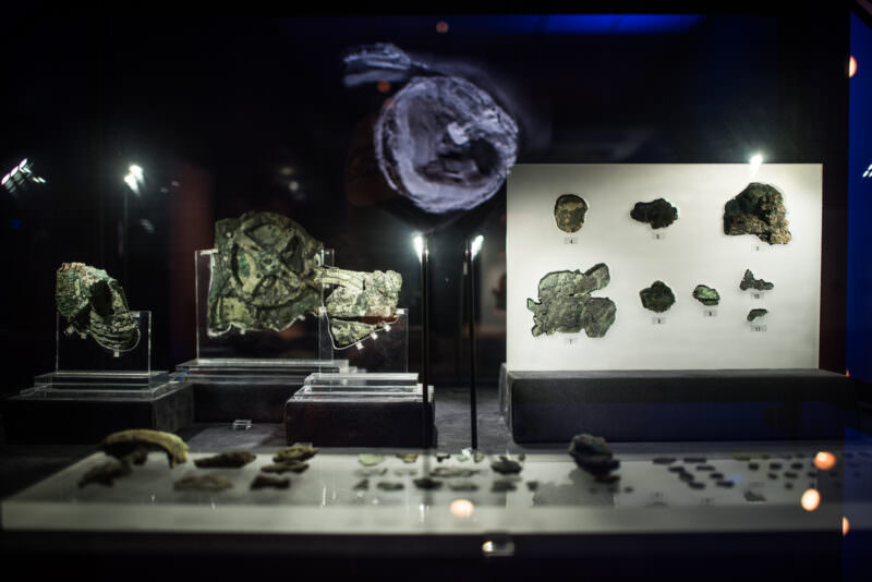 Antikythera Mechanism fragments in display cases with small corroded bronze pieces and a projected X-ray reconstruction above them
